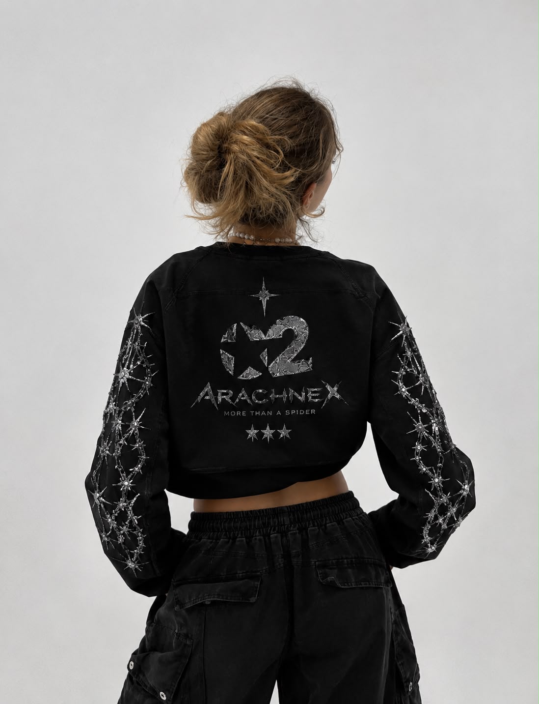 A woman viewed from behind wearing a black cropped long-sleeve top with a large silver graphic and text on the back, plus matching black cargo pants, against a plain light gray studio background.